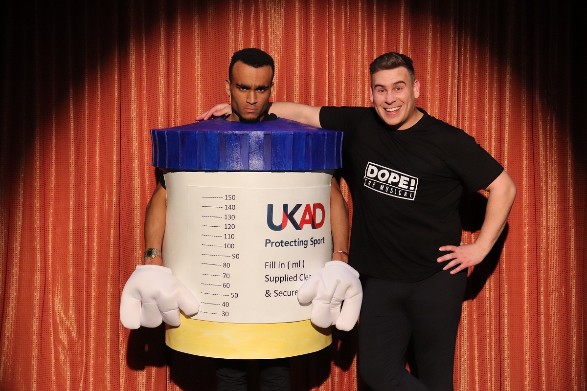 Two people, one wearing a replica urine sample pot